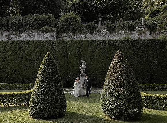 Couple portrait of bride and groom walking hand in hand, veil and bouquet flowing, in a formal garden with hedges and statue