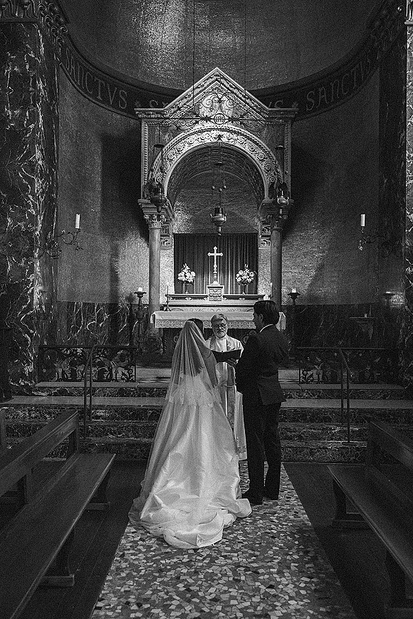 Wedding vows at a church wedding ceremony as bride with long veil and train faces groom and officiant at cross-lit altar with candles