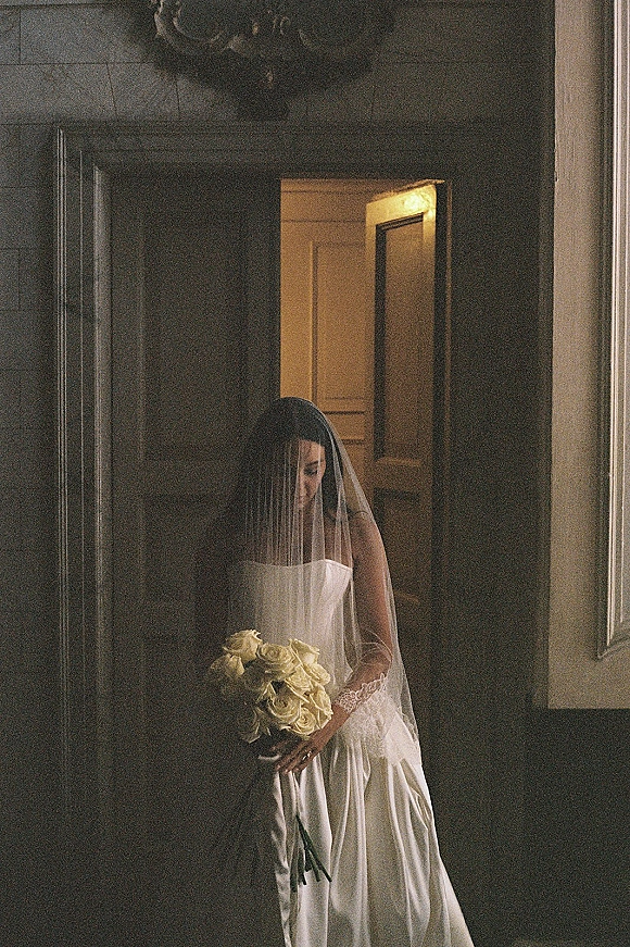 Bridal portrait of a bride holding bouquet in a strapless wedding dress, standing in a doorway with warm light in a shadowy room