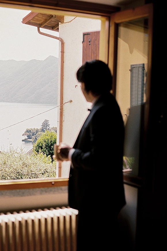 Groom portrait in a black tuxedo and bow tie by a window, looking out at a lake and mountains in soft natural light
