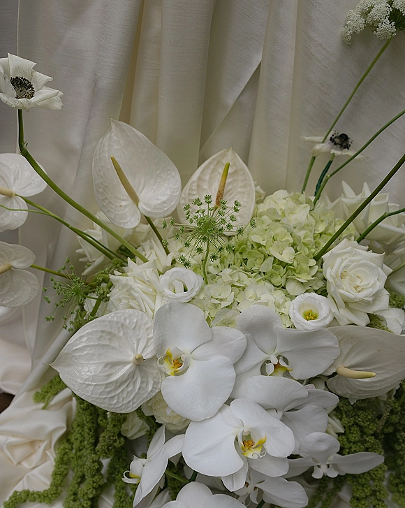 Bridal bouquet featuring white orchids and white anthurium with roses, hydrangea, and greenery stems, tied with satin ribbon on white fabric backdrop