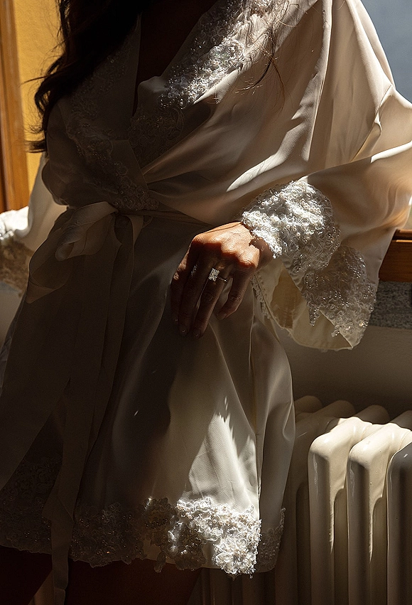 Bridal robe with lace trim and satin belt, styled with lingerie and an engagement ring in soft window light near bed linens
