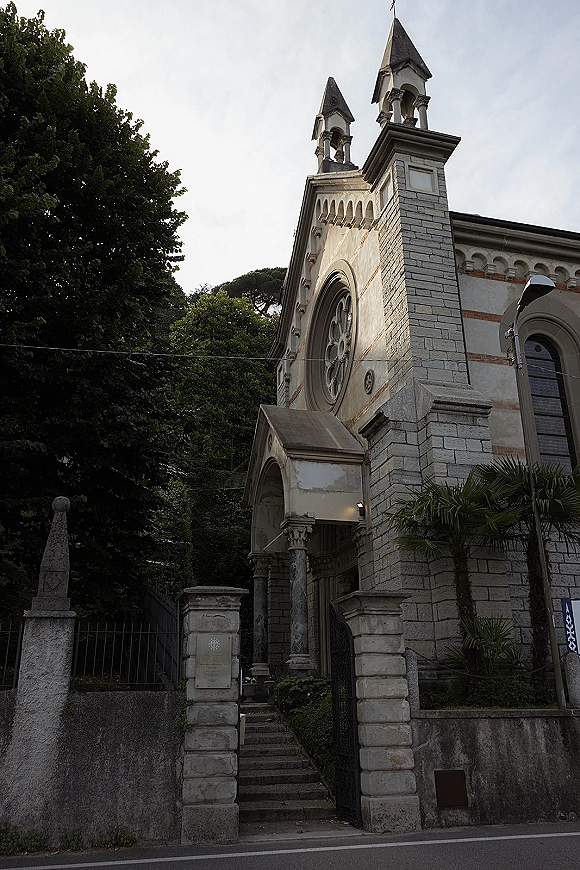 Church exterior with stone facade and twin bell towers, featuring an arched doorway and circular stained glass window above steps and gate