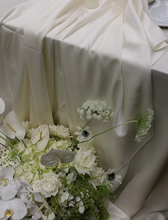 Wedding bouquet of white flowers with roses, orchids, anemones and anthurium resting on draped ivory fabric backdrop