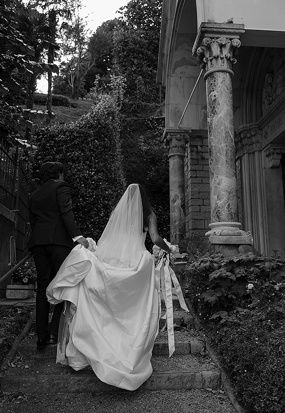 Wedding couple portrait from behind, bride and groom walking away up stone steps, long bridal veil flowing by a columned facade