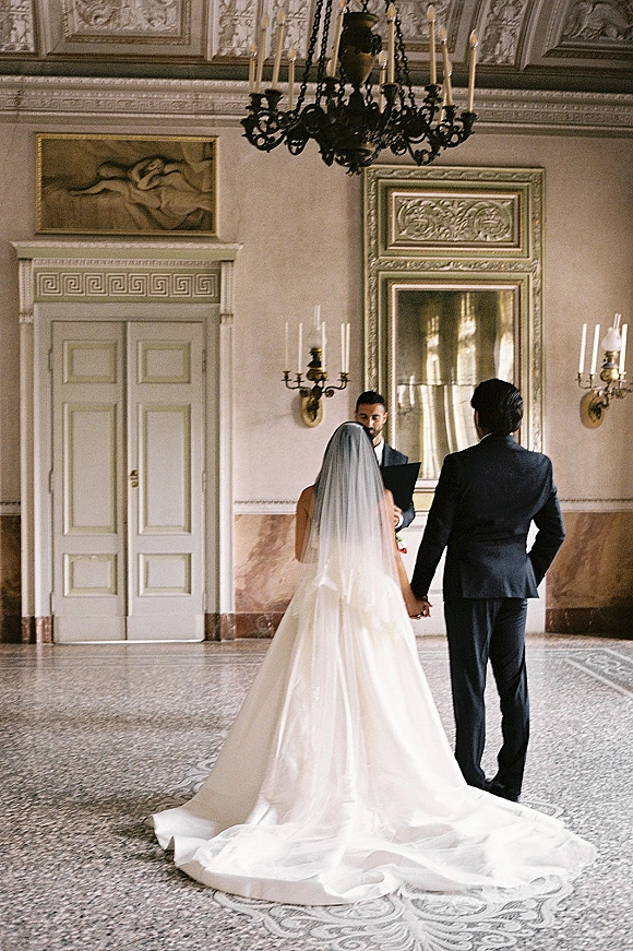 Wedding ceremony moment of couple exchanging vows, holding hands as officiant reads in ornate hall with chandelier and mosaic floor
