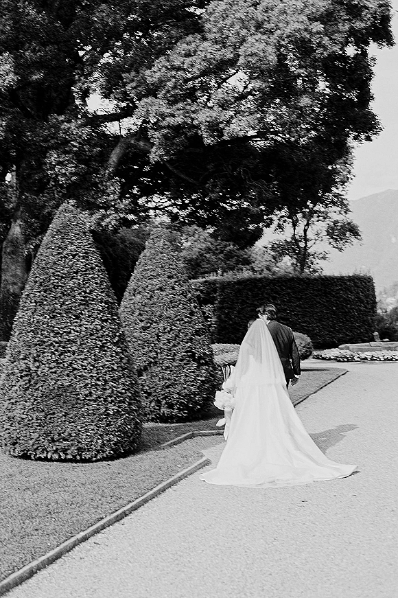 Couple portrait of bride and groom walking away, her long veil and wedding dress train trailing on a gravel path through formal hedges.