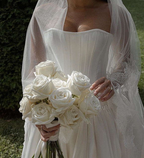 Bridal bouquet of white rose bouquet with ribbon-wrapped handle held by a bride in strapless corset dress and veil on garden lawn