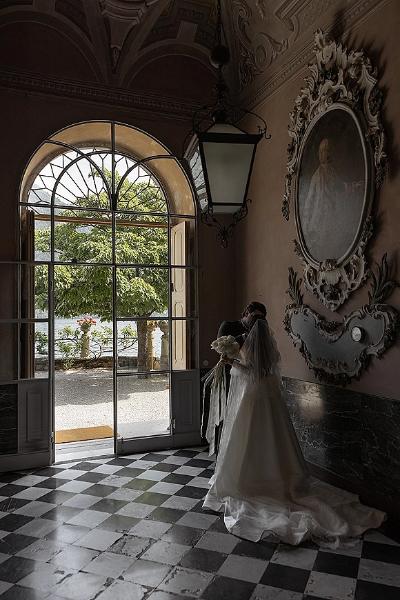 Wedding couple portrait of groom kissing bride’s forehead as she holds a white bouquet with ribbons in a grand arched doorway