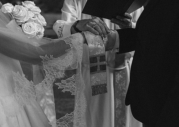 Ceremony moment as bride and groom hands during vows, rings visible beside a white rose bouquet, priest holding a book in church aisle