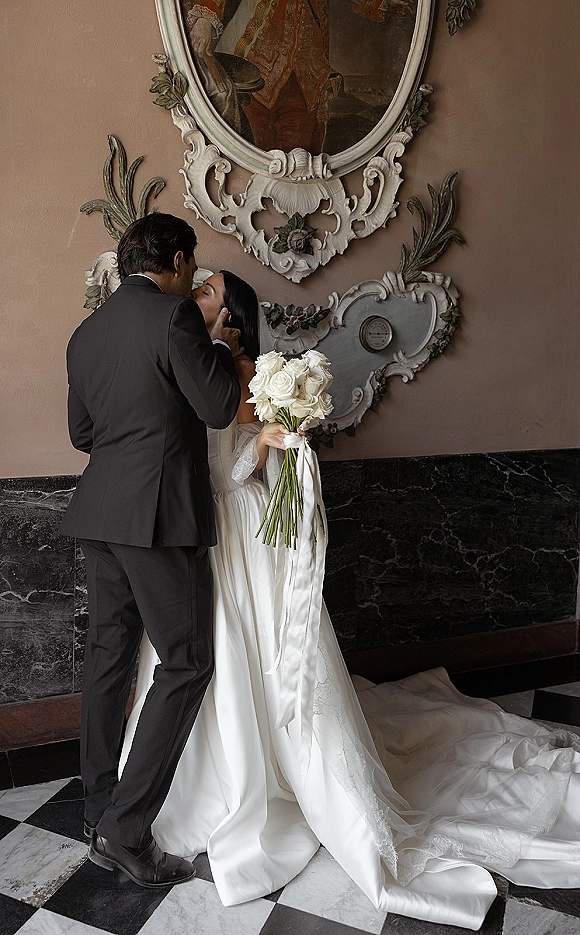 Wedding kiss portrait of bride and groom kissing as she holds white roses with a long ribbon, framed by an ornate mirror and marble wall