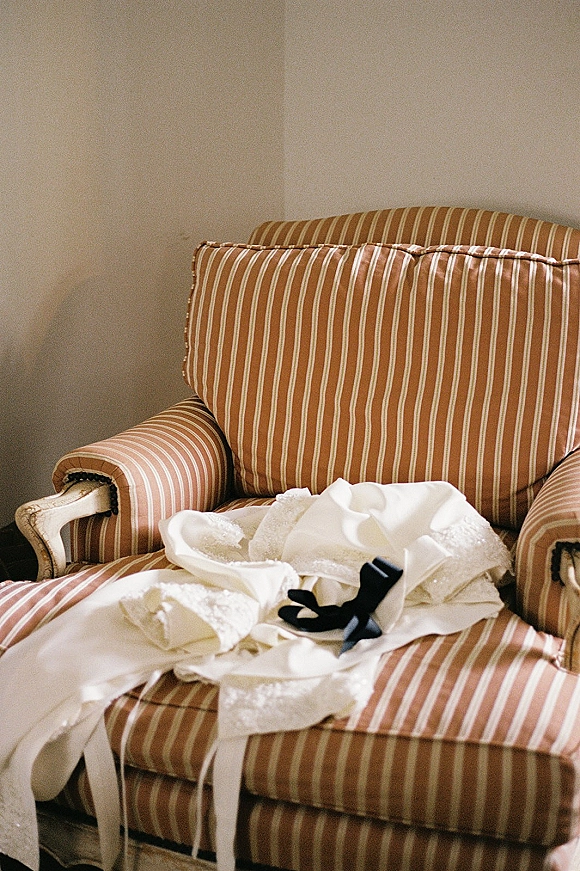 Wedding dress detail with lace trim and a black ribbon bow draped over an upholstered armchair against a plain wall and carpeted floor