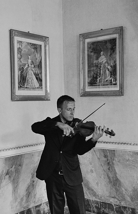 Wedding violinist playing ceremony live music with bow and violin, wearing a suit jacket against a marble gallery wall with framed artwork