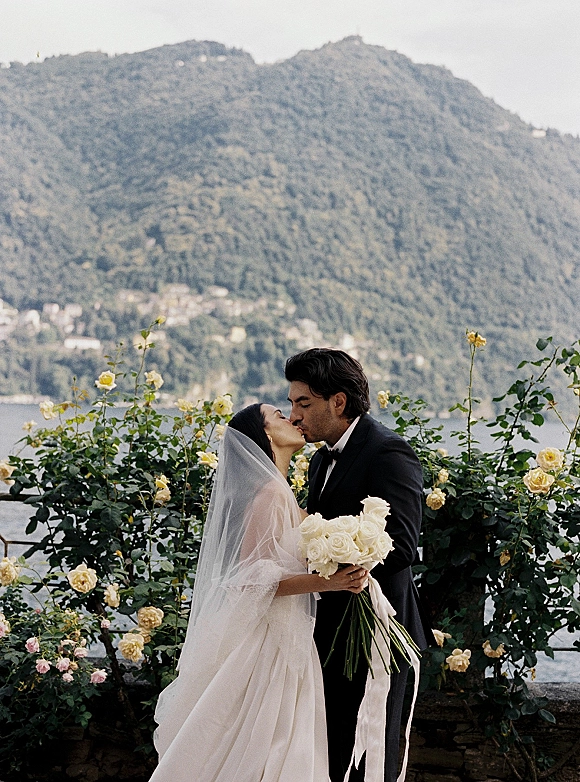 Wedding kiss portrait of bride and groom kissing, her veil and white rose bouquet with ribbon, with mountains and lake behind them
