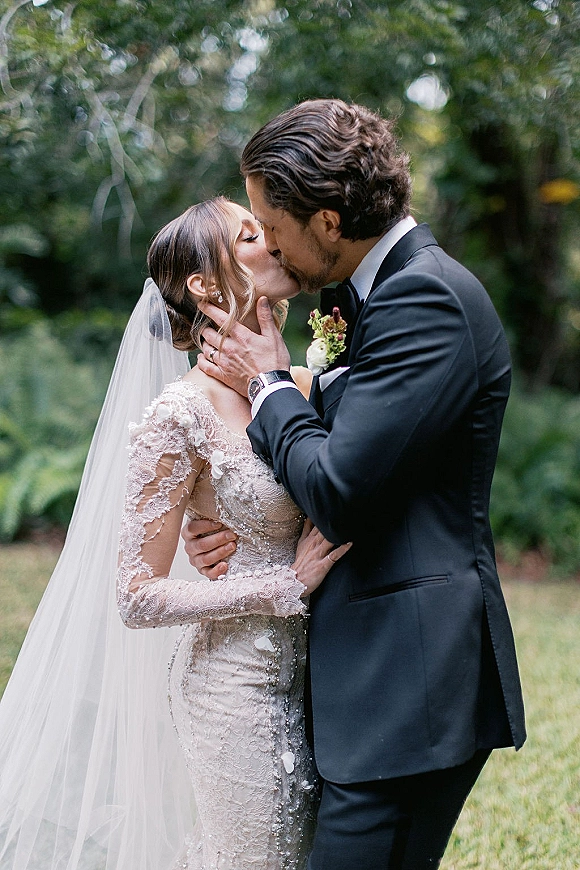 Wedding kiss portrait of bride and groom kissing as he holds her face, lace dress and long veil against a lush green lawn backdrop