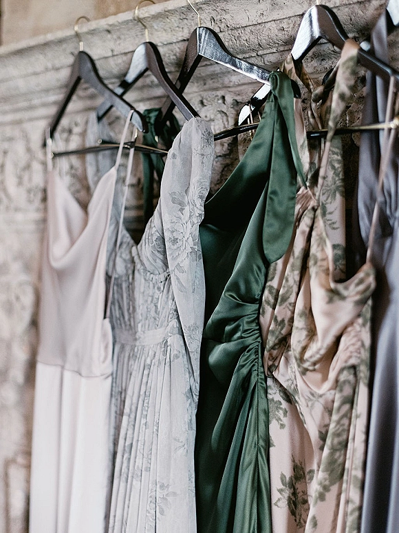 Bridesmaid dresses in sage green hues hang on dress hangers, with satin and floral prints on a rack against a textured wall