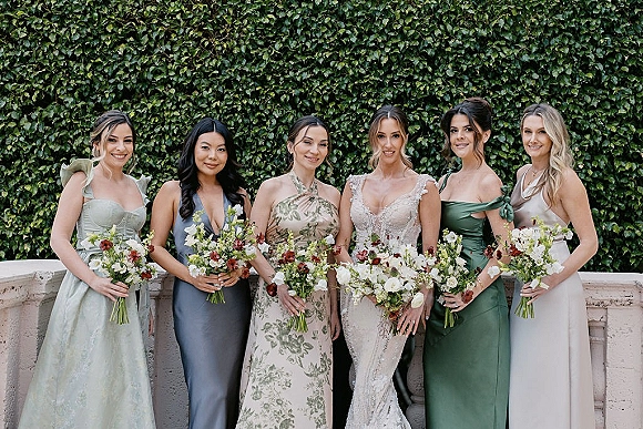 Bridesmaid group photo with bride and bridesmaids holding bouquets in sage green and mismatched dresses by an ivy wall and stone balustrade