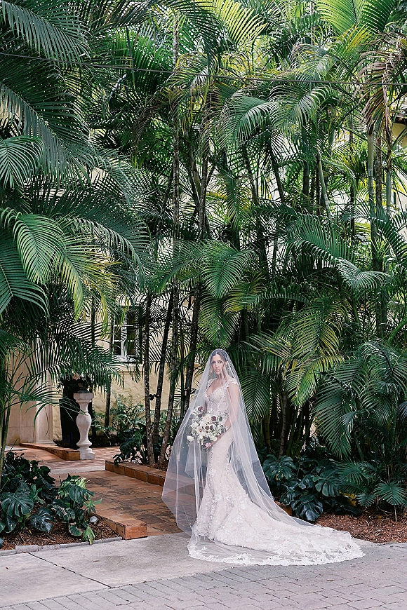 Bridal portrait of a bride with veil in a lace gown holding a bouquet on a brick walkway in a tropical courtyard with palms