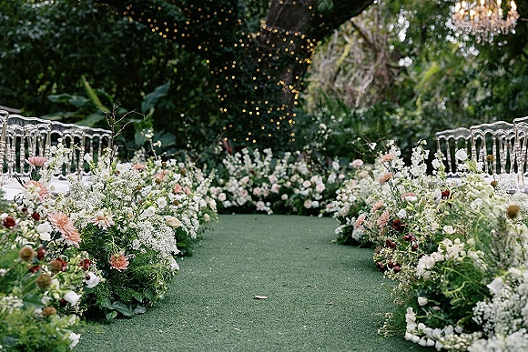 Ceremony aisle decor with ground floral borders of white and blush flowers and greenery, clear chairs under string lights and a chandelier in a garden setting