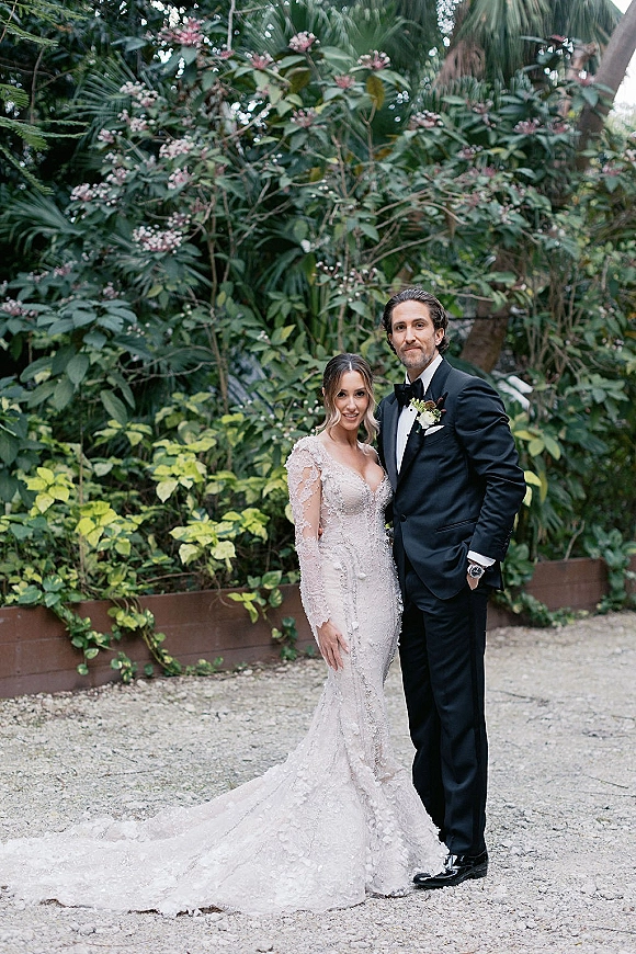 Couple portrait of bride in lace off-the-shoulder gown with long train beside groom in black tuxedo on a lush tropical garden path
