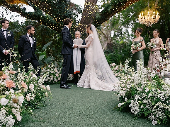 Wedding ceremony with bride and groom exchanging vows beneath a chandelier, string lights, and lush aisle florals in a garden setting