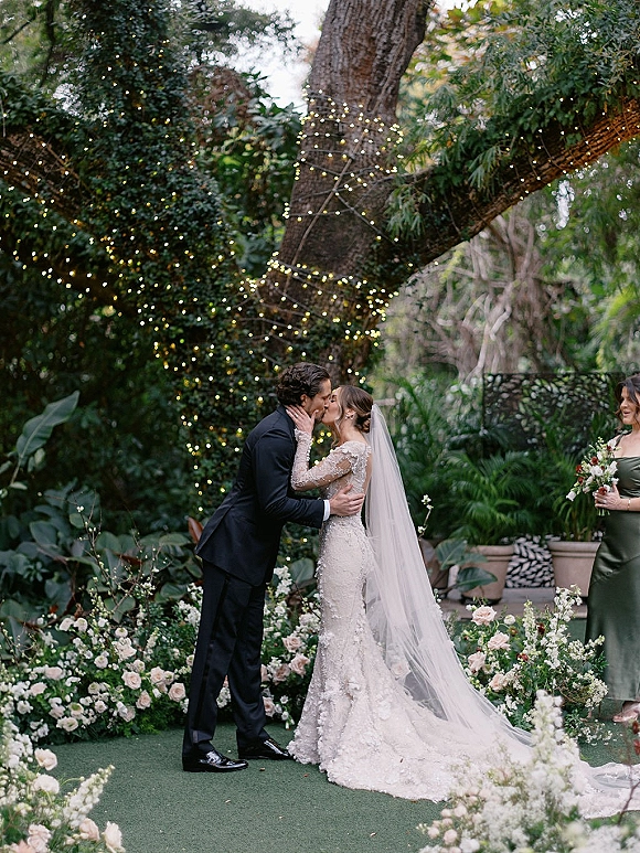 Wedding kiss as bride holds groom’s face, lace dress and long veil flowing down a floral-lined garden aisle under string lights