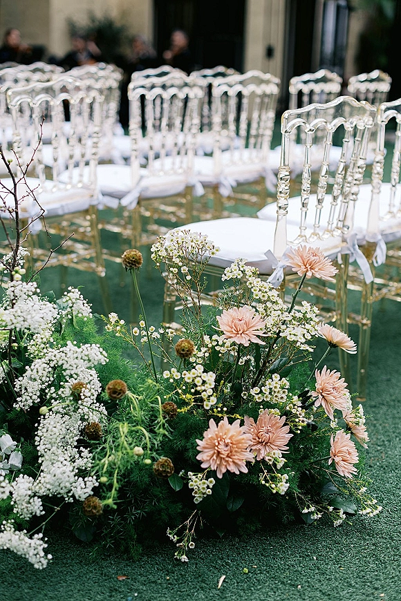 Ceremony aisle decor with clear chiavari ceremony chairs lined in rows, white bows and blush florals with greenery on carpeted floor