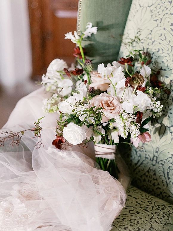 Bridal bouquet of blush and white bouquet blooms with greenery accents, ribbon-wrapped stems, resting on a patterned armchair indoors