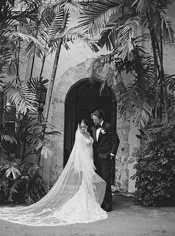 Couple portrait in a black and white wedding portrait style, bride in lace train and long veil with groom in tuxedo under stone arch doorway