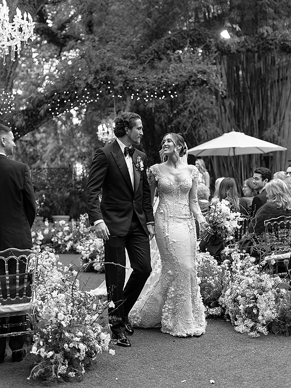 Wedding recessional as bride and groom walk the aisle, her bouquet and veil trailing beside tuxedo, under garden string lights and guests