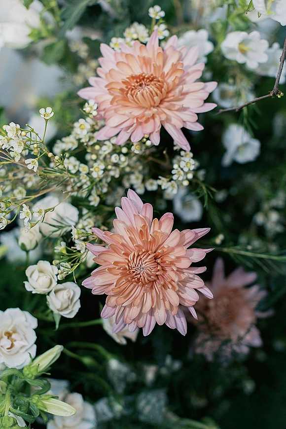 Wedding florals featuring blush wedding flowers with pink chrysanthemums, white roses, baby's breath, and greenery against soft-focus foliage backdrop