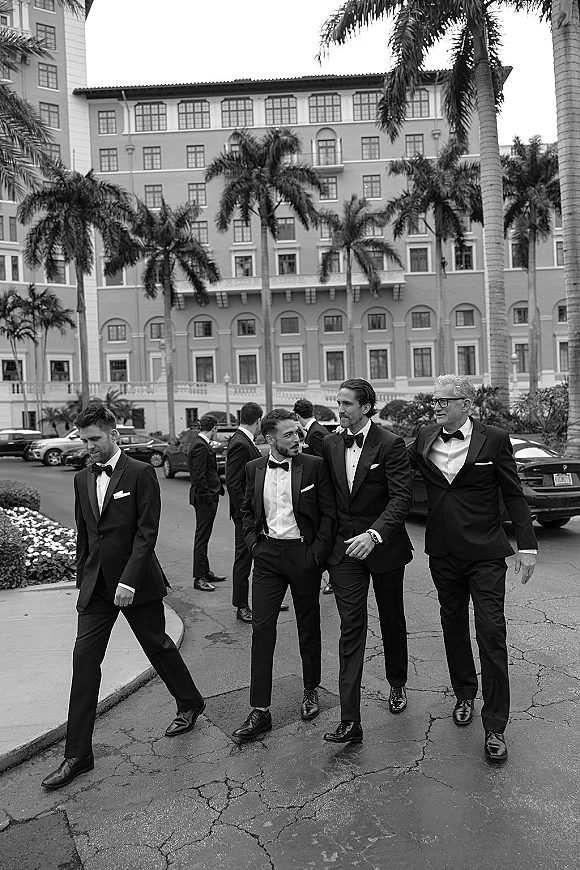 Groomsmen portrait of a groom and groomsmen walking in matching black tuxedos with bow ties and pocket squares by palm-lined hotel driveway