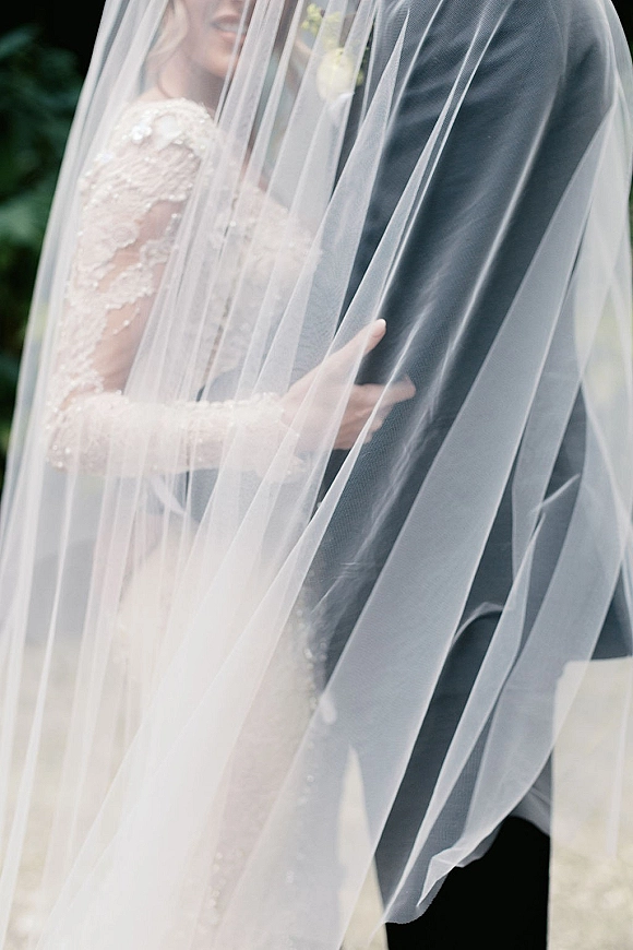 Couple portrait of bride and groom under veil, her lace long-sleeve beaded gown and his boutonniere framed by outdoor greenery