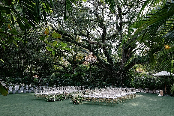 Outdoor ceremony setup with clear chiavari chairs in a semicircle, chandeliers and market lights beneath a large oak tree canopy