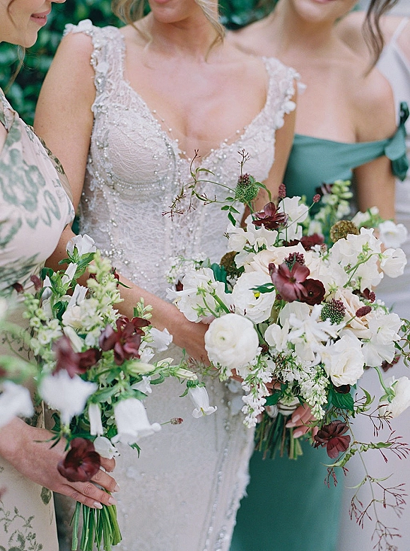 Bridesmaid bouquets of white and burgundy flowers with greenery, held beside a beaded lace bridal bodice against outdoor greenery