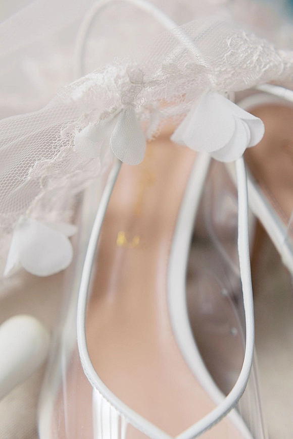 Bridal shoes white wedding heels in a clear shoe box with lace veil and paper petal garland on soft fabric surface