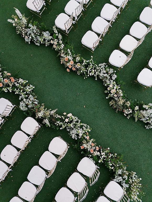 Ceremony aisle decor with a floral aisle runner of white and peach ground flowers and greenery, lined by clear chairs on a green lawn