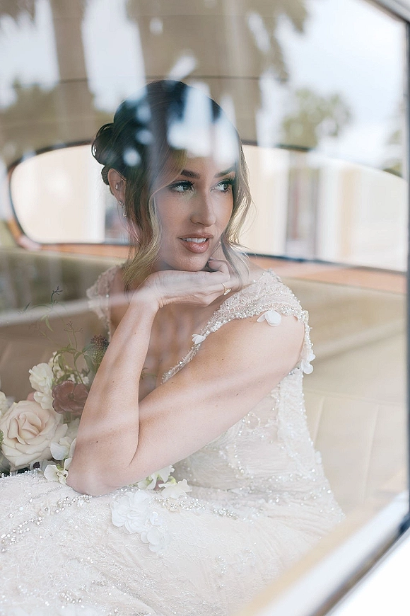 Bridal portrait of a bride in car wearing a beaded wedding dress, resting her chin by the window with bouquet and pearl earrings in daylight reflections