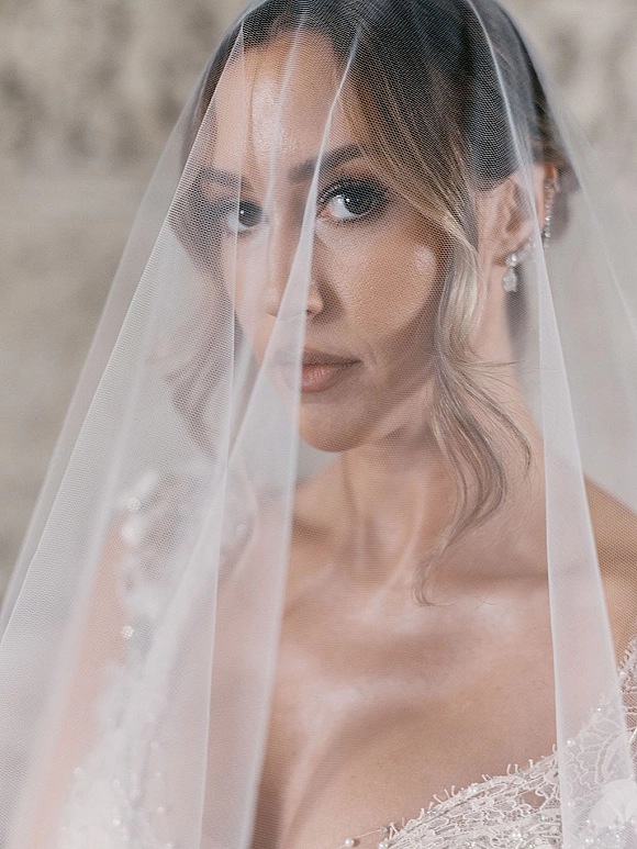 Bridal portrait of a bride with veil over her face, showing soft glam makeup, sparkling drop earrings, and lace dress neckline indoors