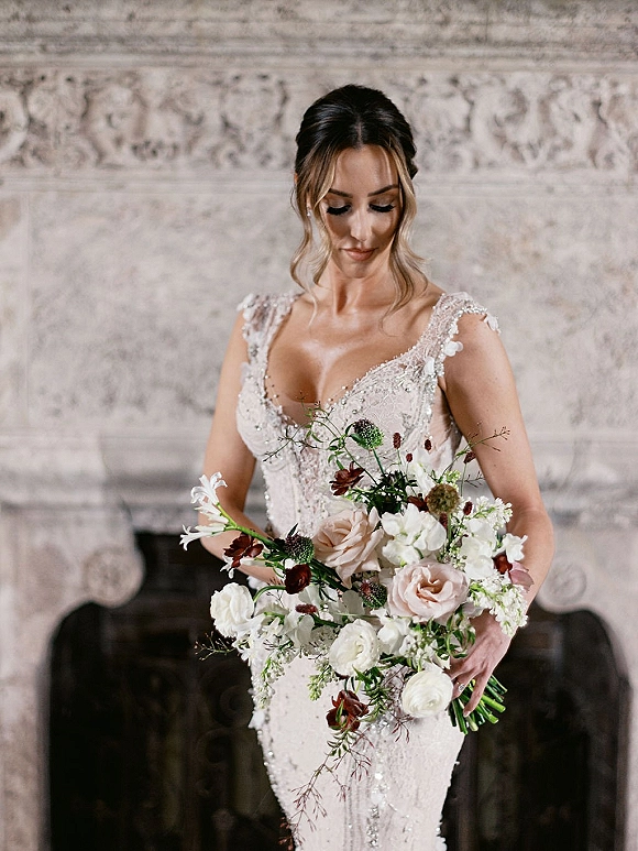 Bridal portrait of a bride holding bouquet of blush and white roses with greenery, in a lace beaded gown by a stone fireplace mantel