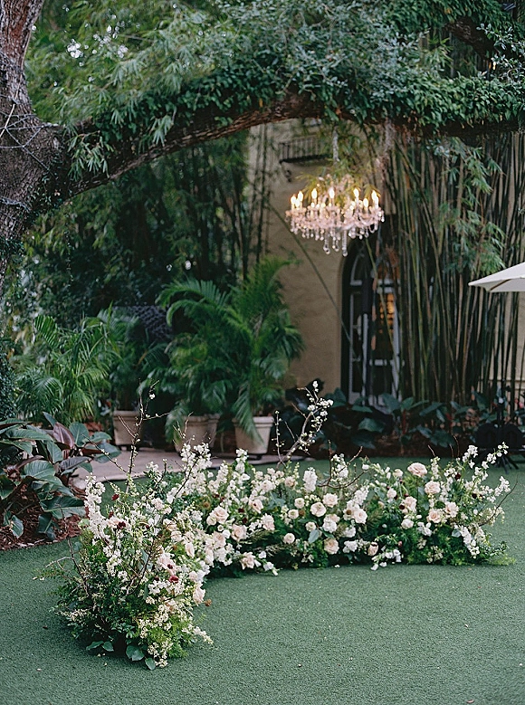 Ceremony aisle decor with ground floral aisle arrangement of white and blush blooms, lush greenery, candles, and a crystal chandelier in a garden courtyard