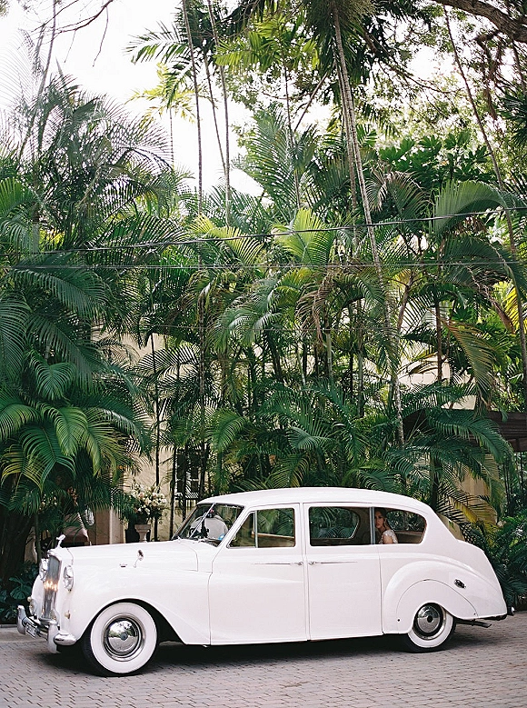 Wedding getaway car with a bride in her bridal gown peeking from a vintage white car window by palm trees on a driveway