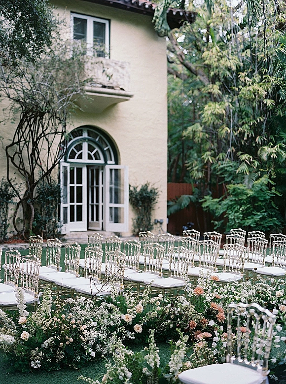Outdoor ceremony setup with clear chiavari chairs and a rose-lined aisle on a garden lawn, framed by trees and a stucco building with arched doorway