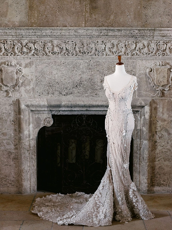 Wedding dress in lace wedding dress style on a mannequin, featuring beaded floral appliques and a long train by a stone fireplace