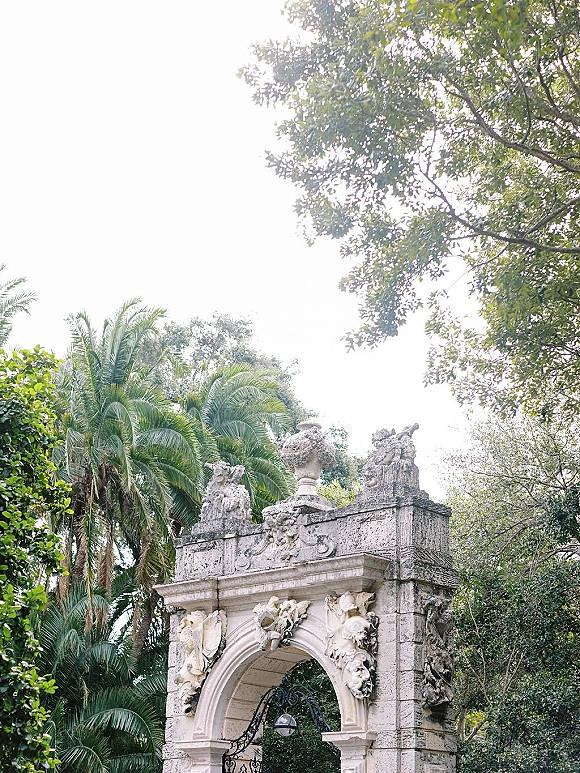 Garden archway with carved stone reliefs and statues, framing a wrought iron gate amid palm trees and lush garden foliage under sky