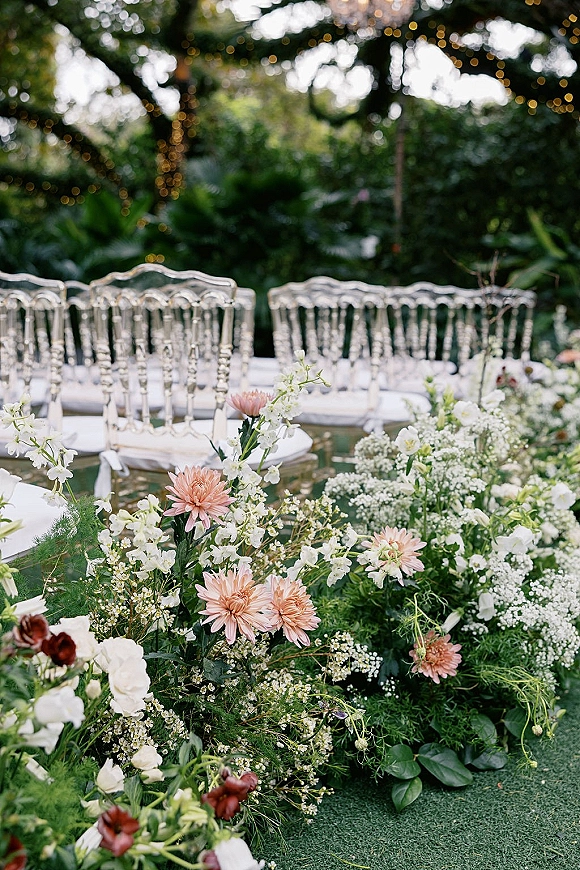 Ceremony aisle decor with floral aisle arrangement of blush and white flowers and greenery beside clear acrylic chairs under string lights in a garden