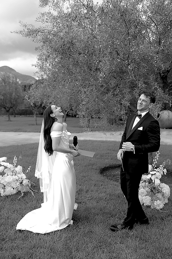 Ceremony moment as bride reads wedding vows into a microphone, groom in black tux reacts on a mountain lawn under cloudy sky