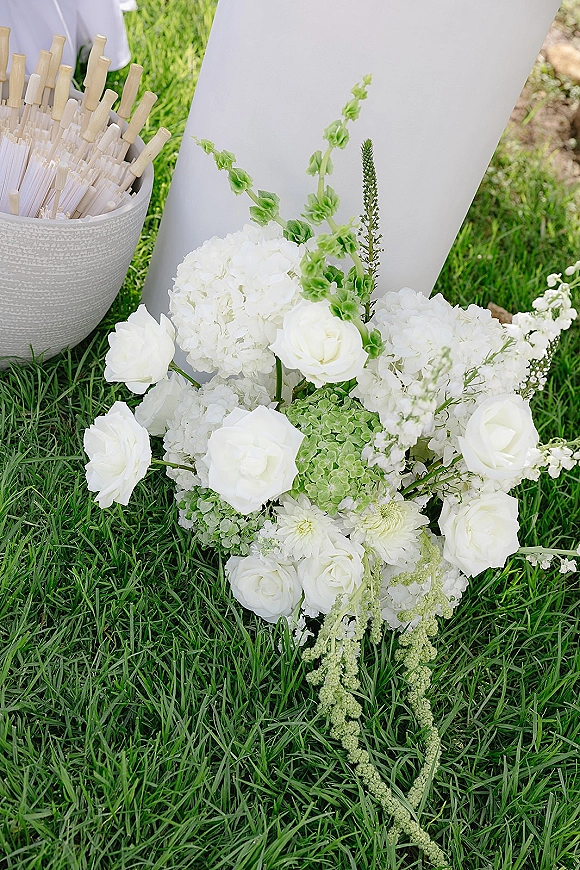 Wedding welcome sign with white hydrangea and rose arrangement at the base, bubble wands in a bowl beside it on grass lawn
