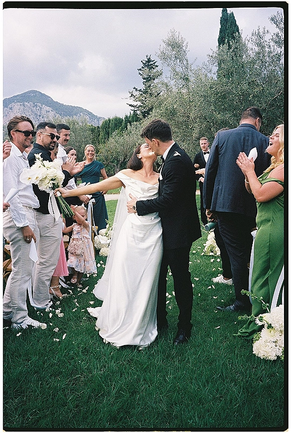 Wedding kiss as newlyweds walk the aisle, bride in off-the-shoulder gown with white bouquet amid rose petals, mountains behind guests