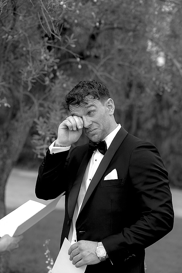 Groom portrait of an emotional groom wiping tears while reading paper notes in a black tuxedo, bow tie, and watch outdoors by trees